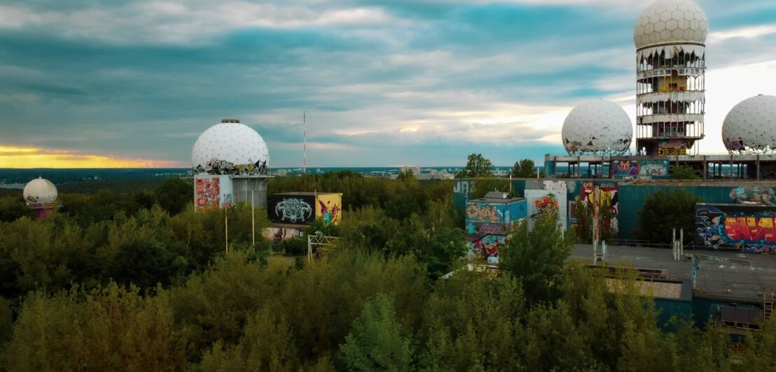 Verlassene Radarkuppeln mit Graffiti auf Gebäuden, umgeben von dichtem Grün unter bewölktem Himmel.