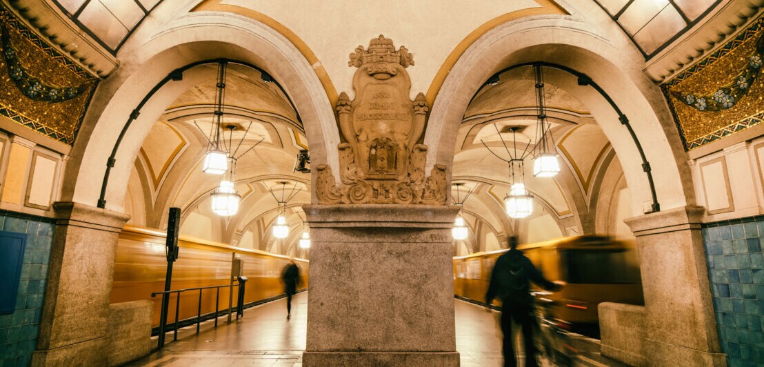 Historische U-Bahn-Station mit großen Bögen, verzierten Säulen und Pendelleuchten, zwei Personen in Bewegung.