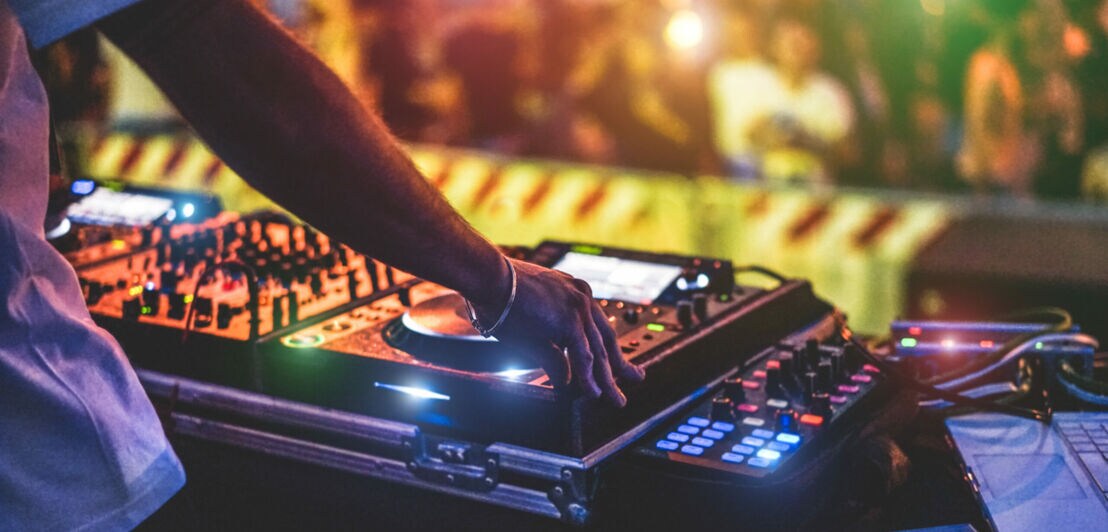 Ein DJ steht vor seinem Mischpult, im Hintergrund ist verschwommen Publikum zu sehen.