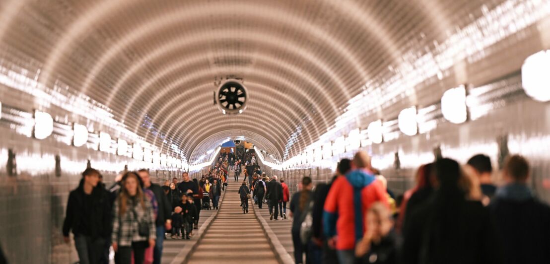 Personen in einem langen, beleuchteten Fußgängertunnel mit gewölbter Decke und weißen Fliesen.