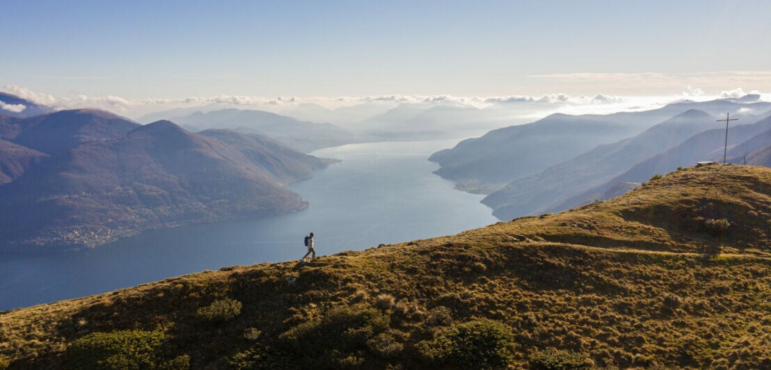 Panoramaaufnahme von einem Mann mit Rucksack auf einem Berg und dem Lago Maggiore im Hintergrund.