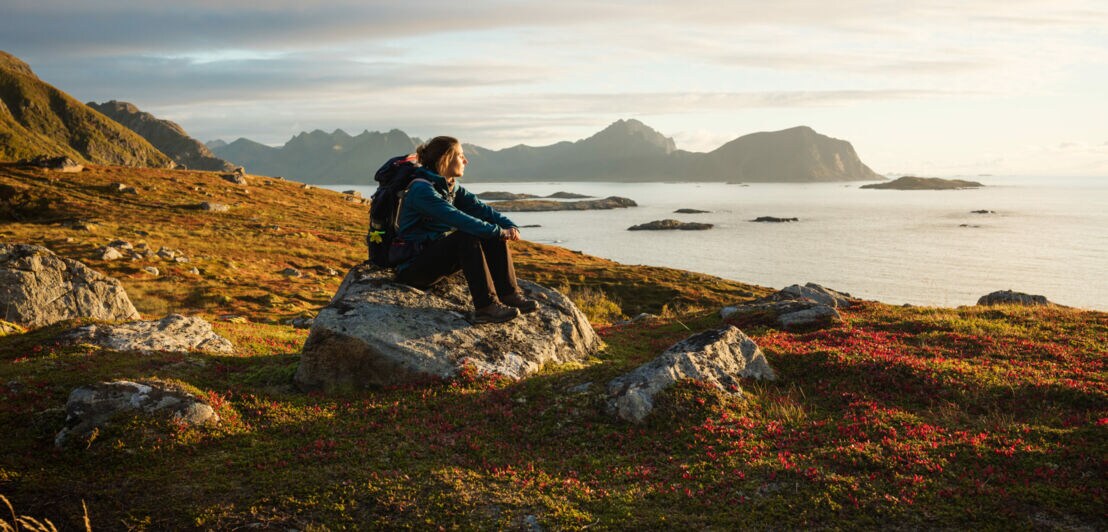 Eine Frau mit Wanderausrüstung, die bei Sonnenuntergang auf einem großen Stein sitzt und aufs Wasser blickt, im Hintergrund Berge.