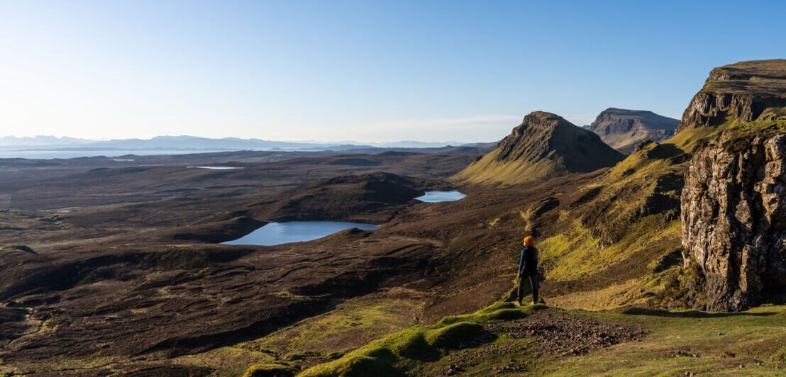Eine Person steht in einer Landschaft aus Wiesen, Hügeln und Seen in Schottland.