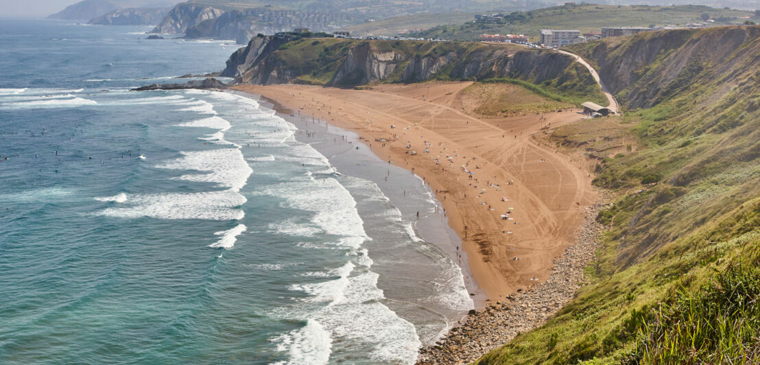 Langer Sandstrand mit Wellen und Menschen, umgeben von steilen Klippen und grünen Hügeln unter bewölktem Himmel.