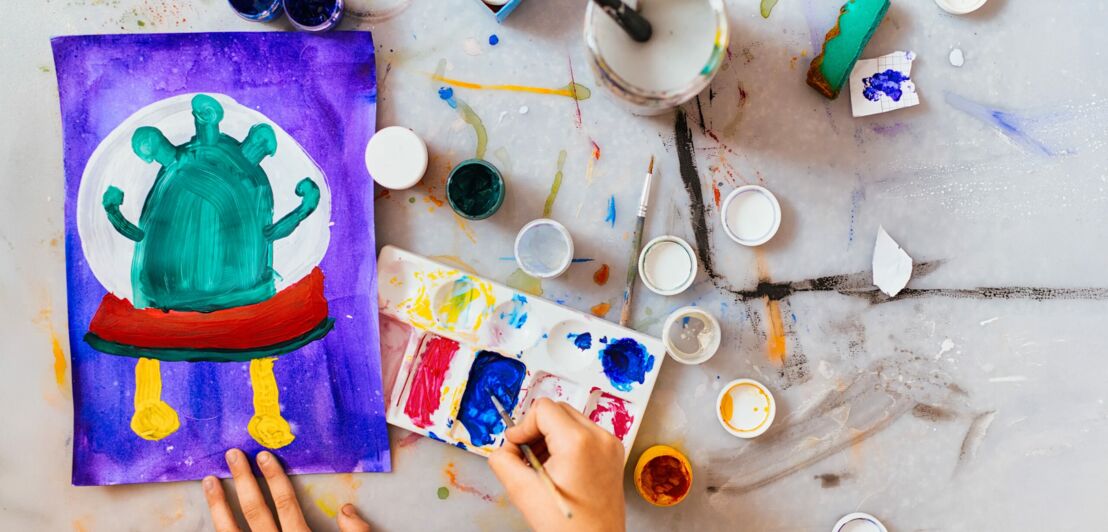 Kind malt eine grüne, fantasievolle Kreatur mit gelben Beinen und rotem Gürtel auf lila Hintergrund mit Wasserfarben und Pinseln auf einem Tisch