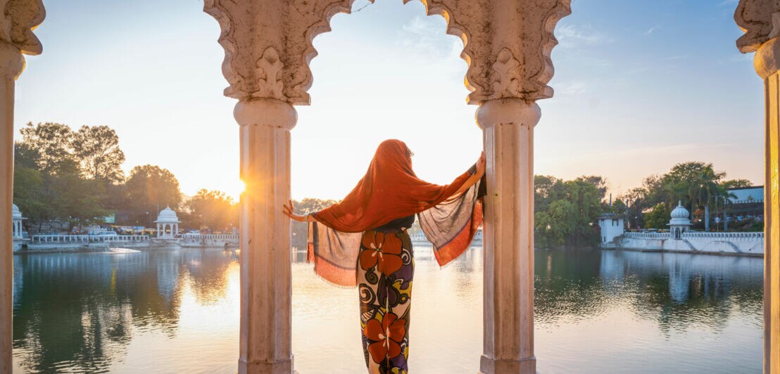 Eine Frau in traditioneller indischer Kleidung steht in einem Pavillon und blickt aufs Wasser.