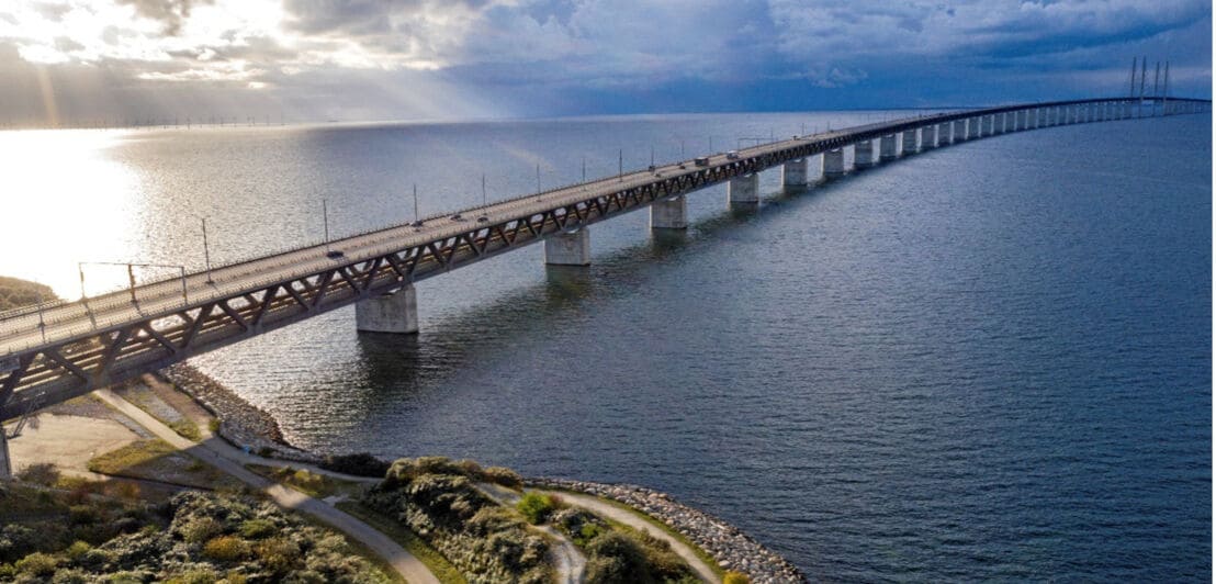 Lange Brücke über ruhiges Meer mit bewölktem Himmel und Sonnenlicht, das durch die Wolken scheint.