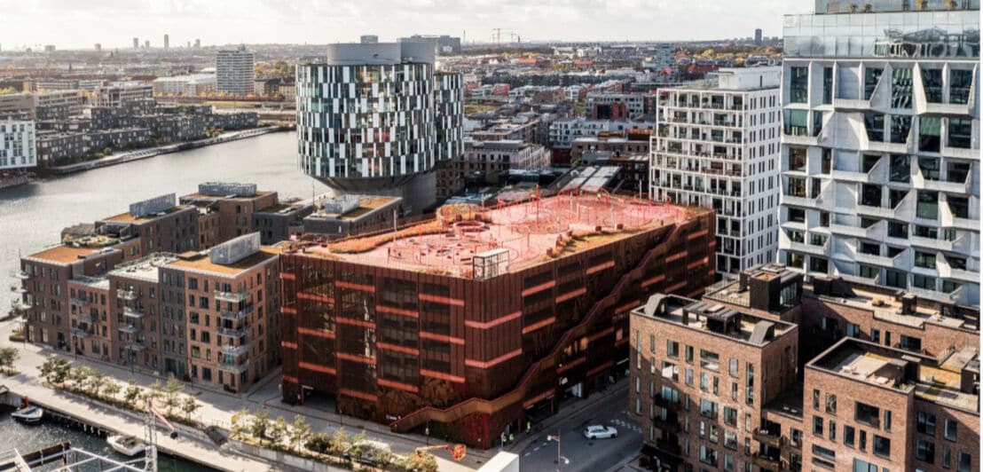 Städtische Architektur mit modernen Wohn- und Bürogebäuden entlang eines Kanals, darunter ein auffälliges rotes Gebäude mit Dachterrasse und ein rundes Gebäude mit gemusterten Fassaden.