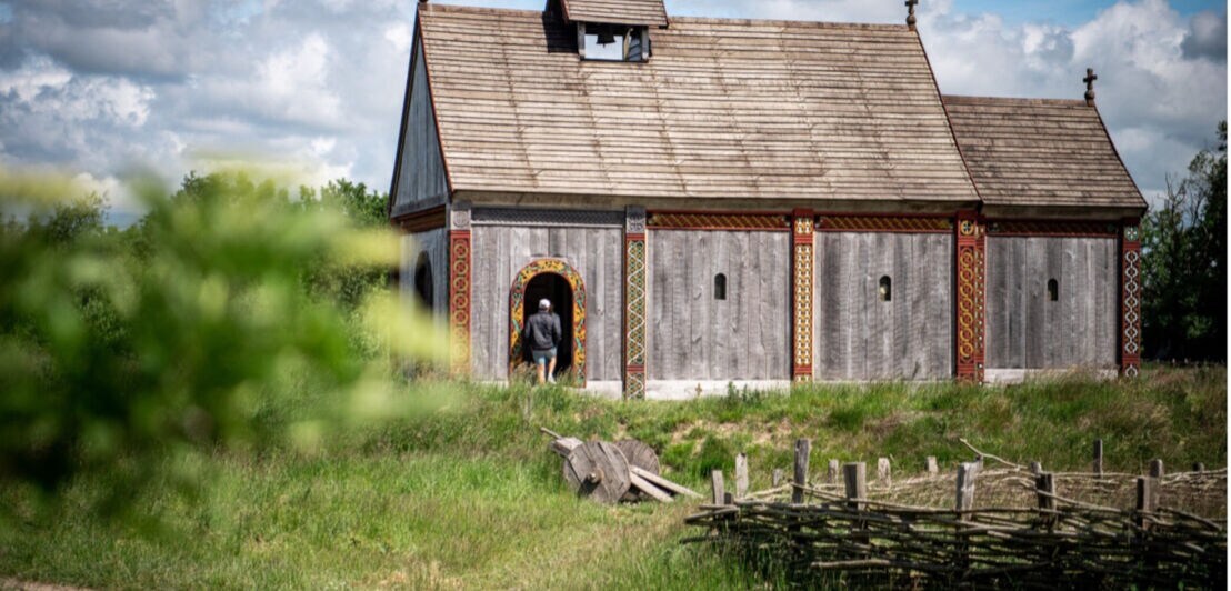 Holzgebäude mit verzierten Tür- und Fensterrahmen, umgeben von grünem Gras und einem Weidenzaun, Person betritt das Gebäude.
