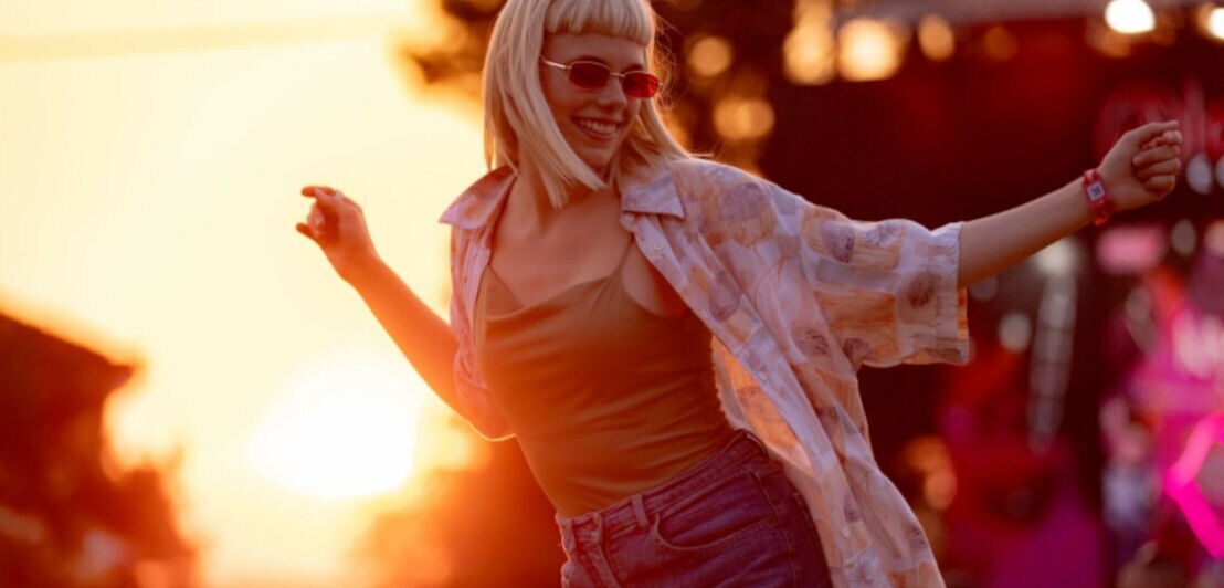 Eine stylisch gekleidete Frau in Shorts mit Sonnenbrille und kurzem Pony tanzt auf einem Festivalgelände im warmen Licht bei Sonnenuntergang.