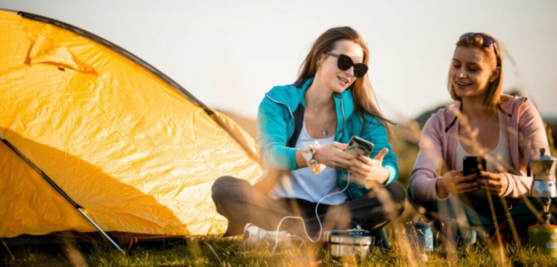 Zwei junge Frauen sitzen mit Smartphones vor einem Zelt auf einem Feld, vor ihnen stehen ein Campingkocher und andere Küchenutensilien.