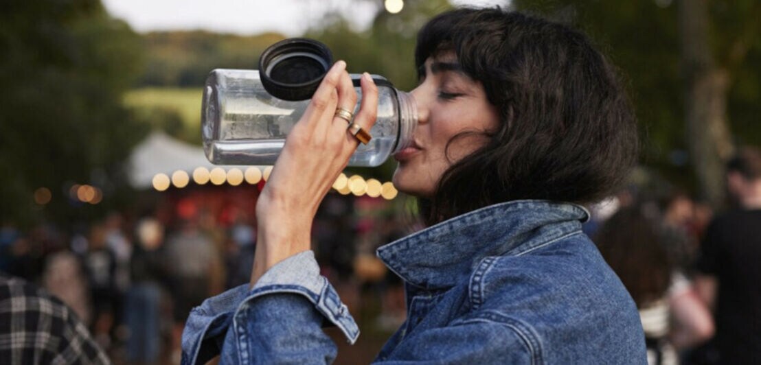 Eine junge Frau in Jeansjacke trinkt aus einer großen Wasserflasche auf einem Festivalgelände.