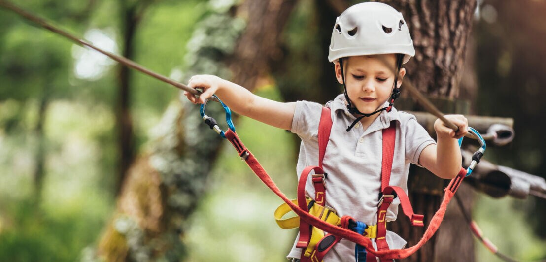 Kind mit weißem Helm und Kletterausrüstung hält sich an Seilen in einem Hochseilgarten fest