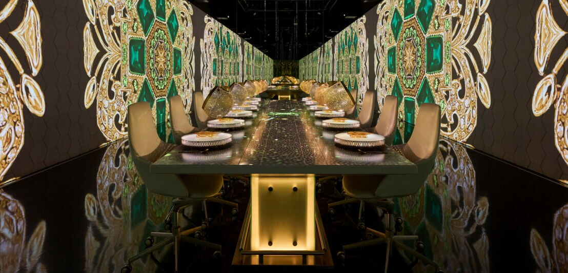 Langer Esstisch mit gedeckten Plätzen in einem Raum mit großen, dekorativen Wandmustern in Grün und Gold