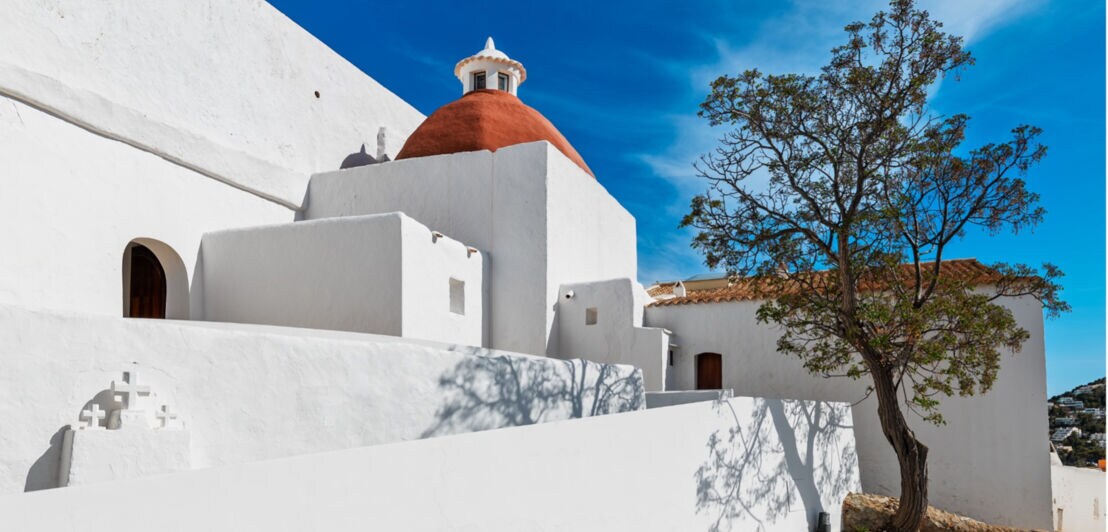 Weiße Gebäude mit roter Kuppel und Baum vor blauem Himmel in mediterraner Landschaft