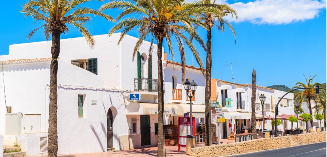 Weiße Gebäude mit Arkaden und Balkonen an einer sonnigen Straße mit Palmen unter blauem Himmel