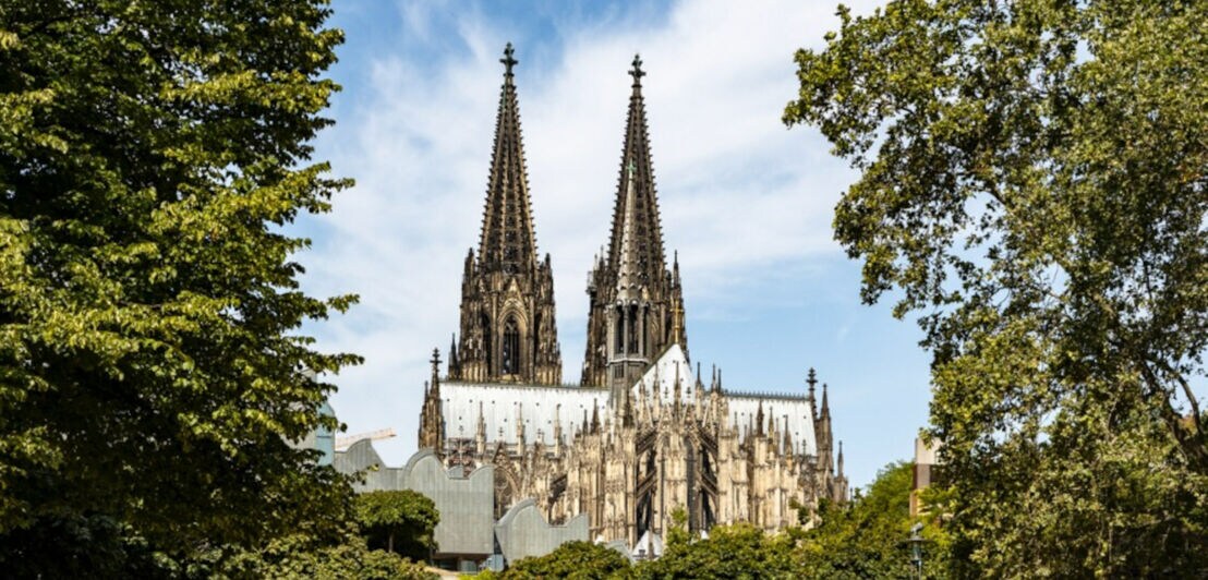 Kölner Dom mit zwei hohen gotischen Türmen, umgeben von Bäumen und blauem Himmel