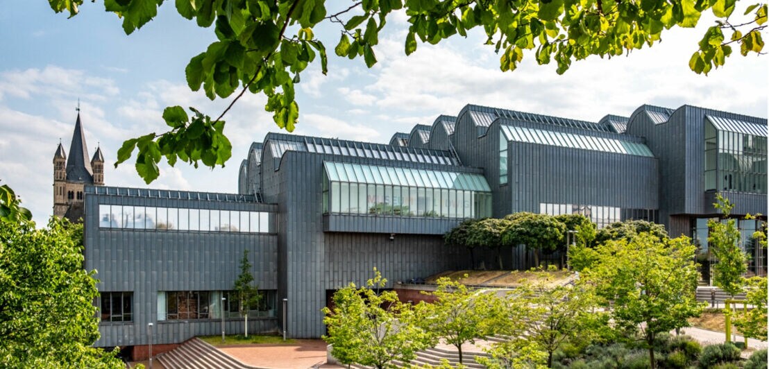 Modernes, mehrteiliges Gebäude mit Glasdächern und Metallfassade, umgeben von Bäumen und Grünflächen, im Hintergrund Kirchturm