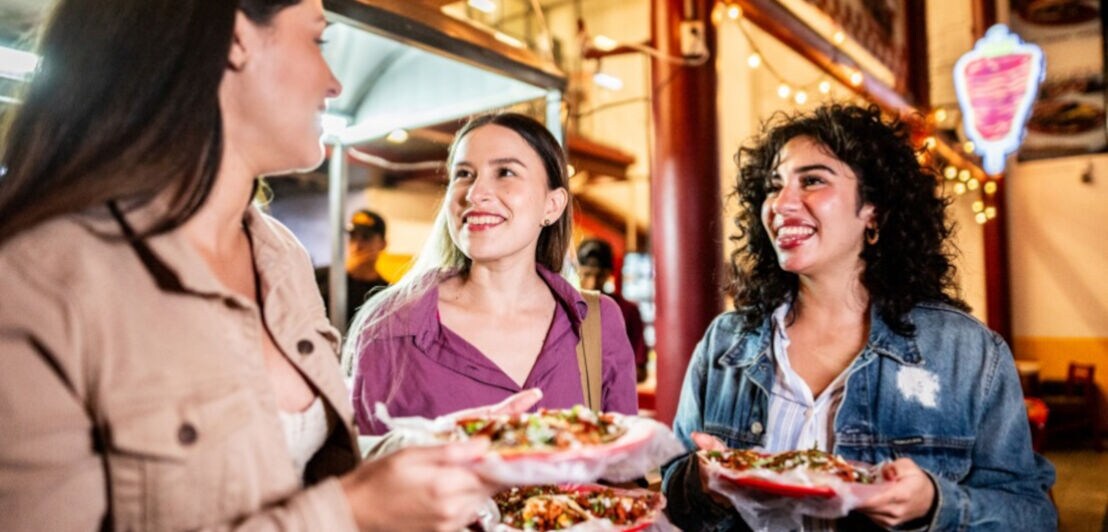 Drei junge Frauen mit Taco-Gerichten in den Händen auf einem beleuchteten Street-Food-Markt bei Nacht.