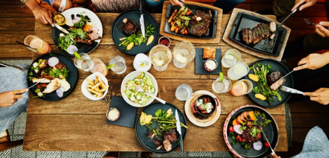 Aufsicht eines Holztisches mit unterschiedlichen Fleischgerichten und Getränken, am Rande die Hände mehrerer Personen mit Besteck.