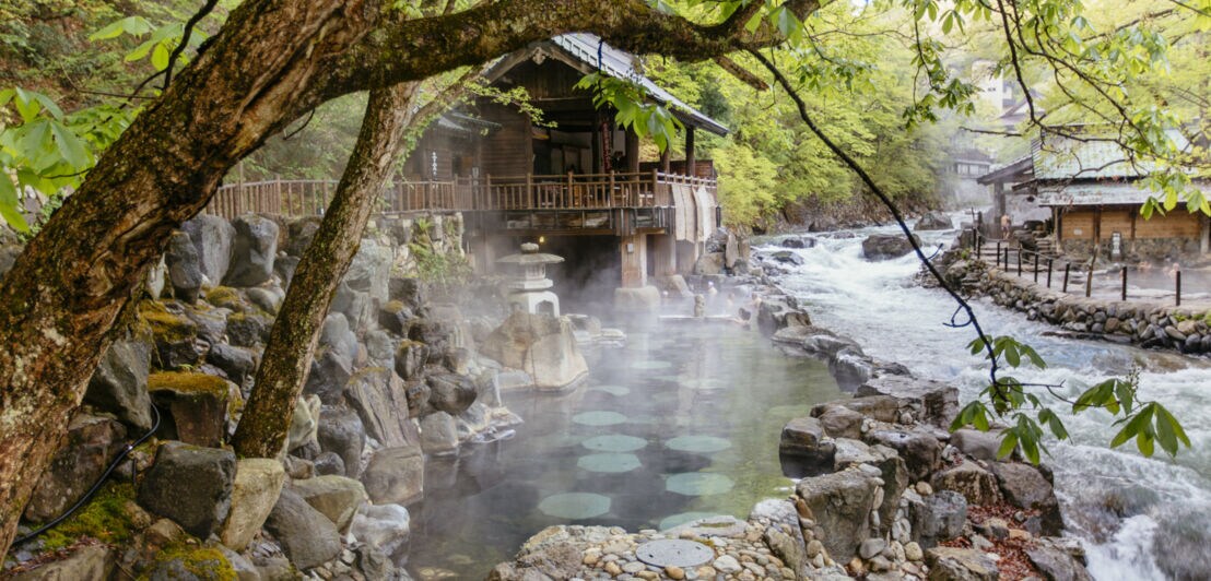 Thermalbad in einem Felsen an einem Fluss mit Holzhütten, umgeben von grünen Bäumen.