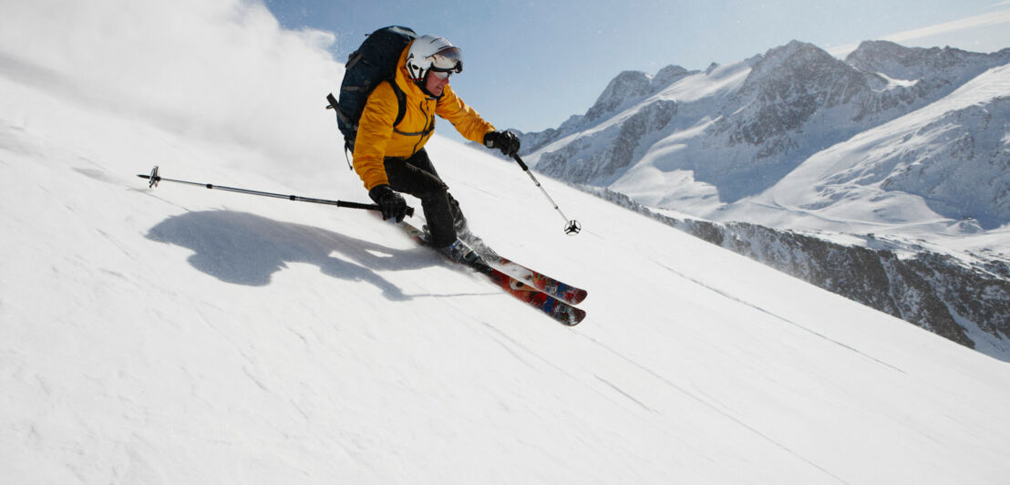 Ein Skifahrer fährt einen steilen Abhang hinunter, im Hintergrund schneebedeckte Berge.