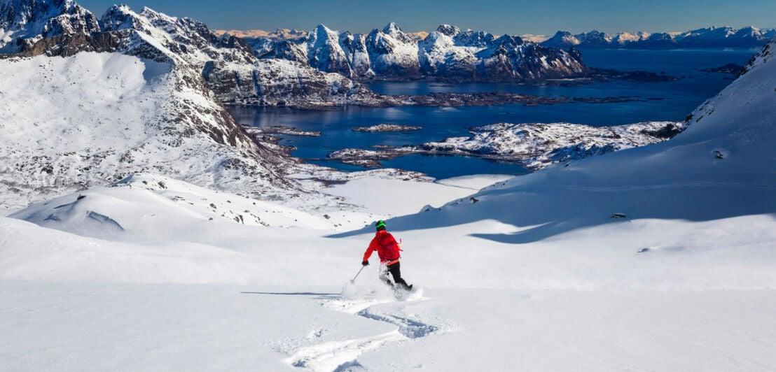 Ein Skifahrer in einer roten Jacke fährt durch den Schnee, im Hintergrund sind Berge.