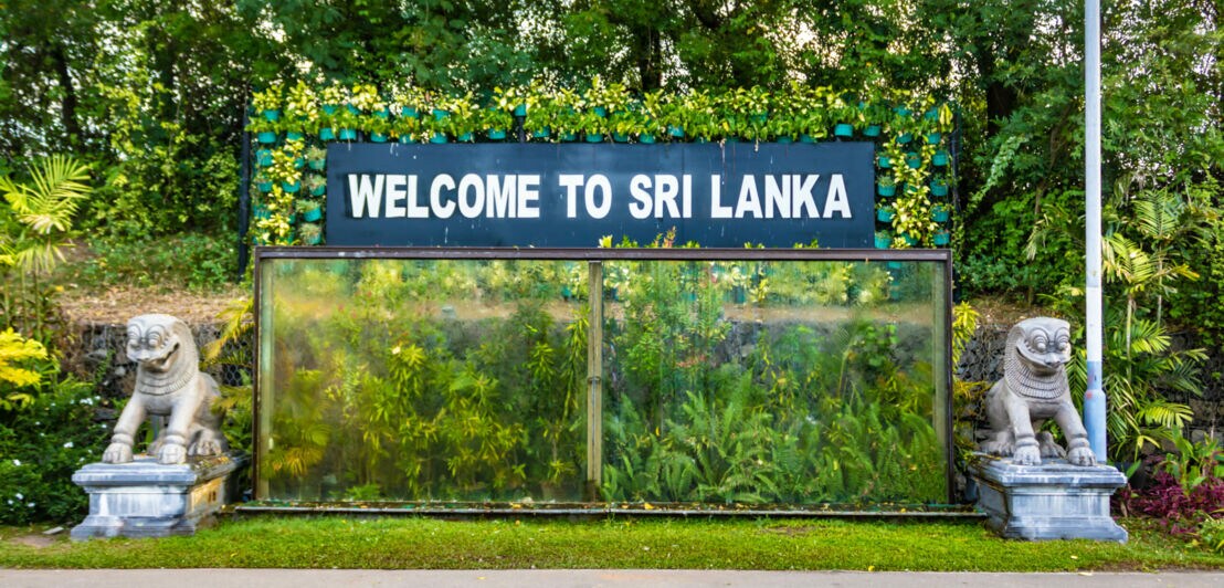 Grüne Pflanzen, vor denen auf einem Schild Welcome to Sri Lanka steht, rechts und links davon eine Skulptur.