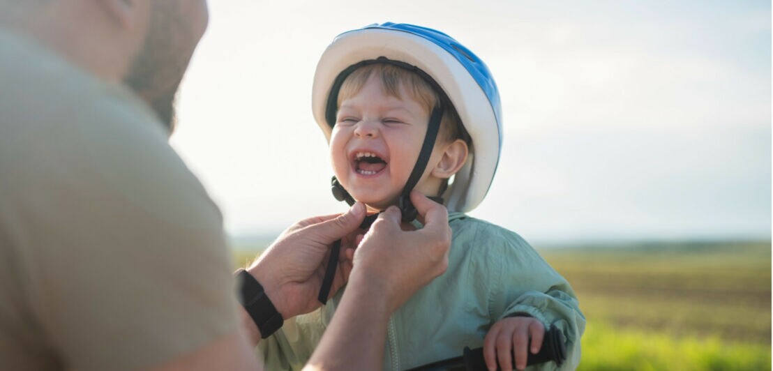 Kind mit blau-weißem Fahrradhelm hält Lenker eines Fahrrads, während eine erwachsene Person den Helm festzieht