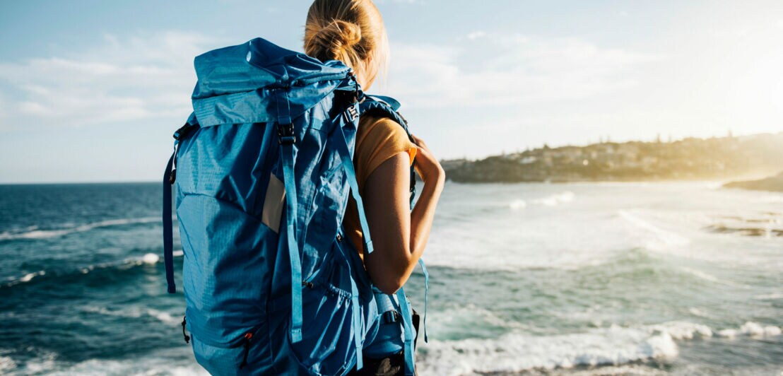 Eine Frau mit einem großen Reiserucksack steht an der Küste und blickt auf das Meer.
