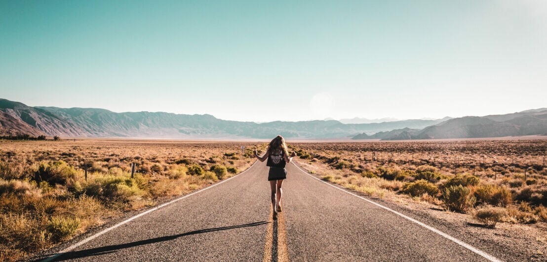 Eine Frau mit Rucksack läuft mitten auf einer leeren Straße, im Hintergrund sind Berge.