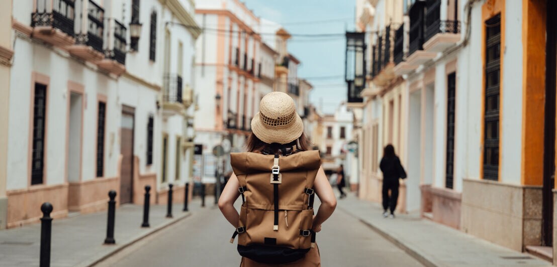 Eine Frau mit einem Rucksack und einem Hut ist von hinten zu sehen. Sie steht in einer Gasse mit hübschen Altbauten.