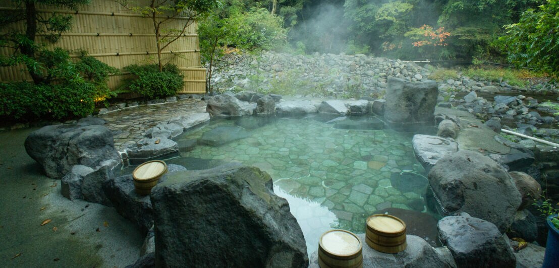 Flaches, dampfendes, von Steinen gerahmtes Thermalbad in einem japanischen Garten, im Vordergrund mehrere Saunakübel aus Holz auf Steinen.