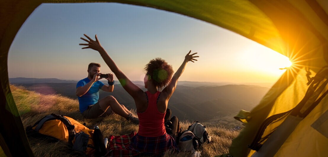 Blick aus einem Zelt auf zwei Personen auf einer Bergwiese bei Sonnenuntergang, eine Person macht ein Foto, die andere hebt die Arme in die Luft