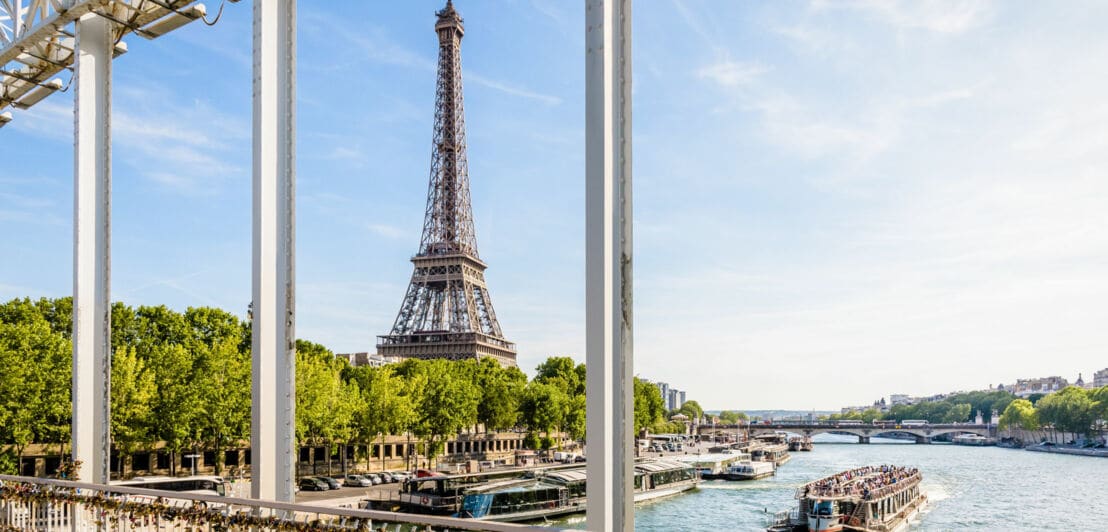 Eiffelturm hinter der Seine, gesehen von einer Brücke mit zahlreichen Liebesschlössern am Geländer, ein Ausflugsboot auf dem Fluss