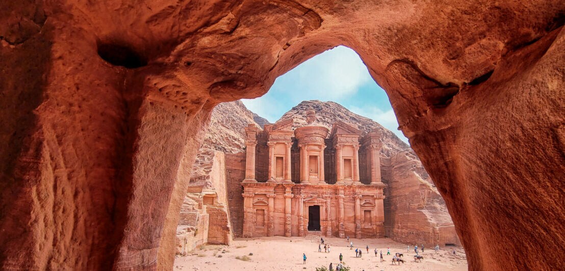 Blick durch eine Felsöffnung auf das antike Kloster Monastery in Petra, Jordanien, mit Touristen im Vordergrund