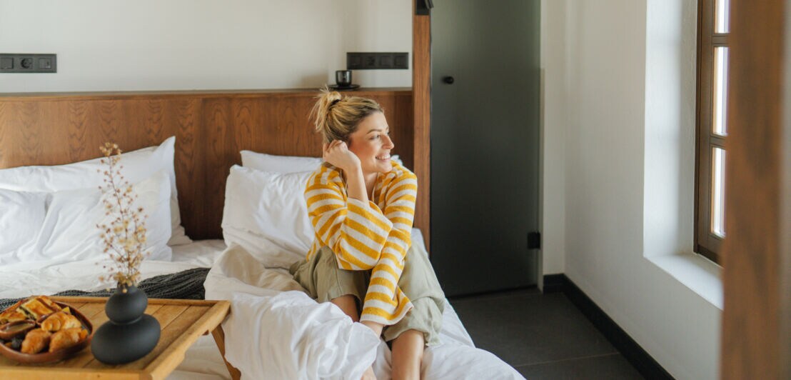 Person sitzt entspannt auf einem Bett mit weißer Bettwäsche in einem hellen Schlafzimmer mit Holzdetails und einem Fenster mit Jalousie