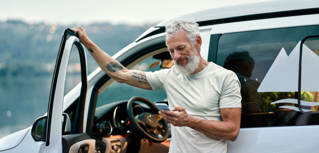 Älterer Mann mit grauen Haaren und Tattoos lehnt an geöffnetem Türrahmen eines weißen Vans und schaut auf sein Smartphone