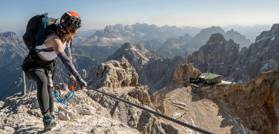 Frau mit Kletterausrüstung auf einem Berg an einem Drahtseil, im Hintergrund Berge.