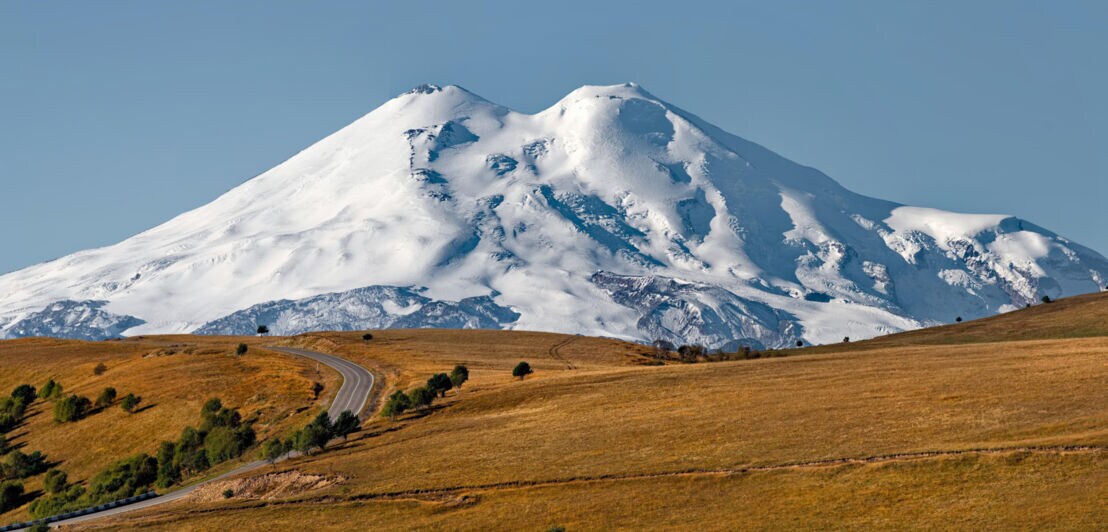 Schneebedeckter Berg mit zwei Gipfeln hinter hügeliger, brauner Landschaft und kurviger Straße unter klarem blauem Himmel