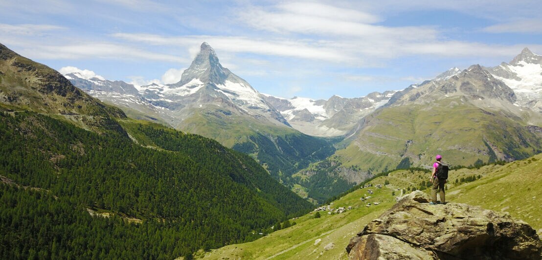 Wanderer mit Rucksack steht auf Felsen und blickt auf das Matterhorn und grüne Alpenlandschaft
