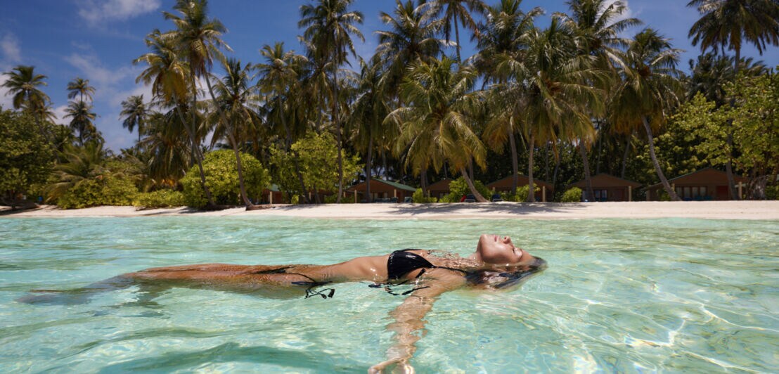 Person schwimmt entspannt auf dem Bauch im klaren, flachen Wasser vor einer tropischen Insel mit Palmen
