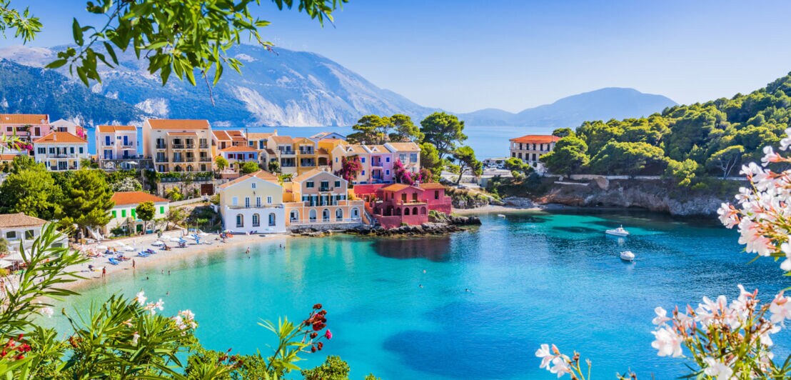 Bunte Häuser am Ufer einer ruhigen Bucht mit türkisfarbenem Wasser, umgeben von grünen Hügeln und Bergen im Hintergrund