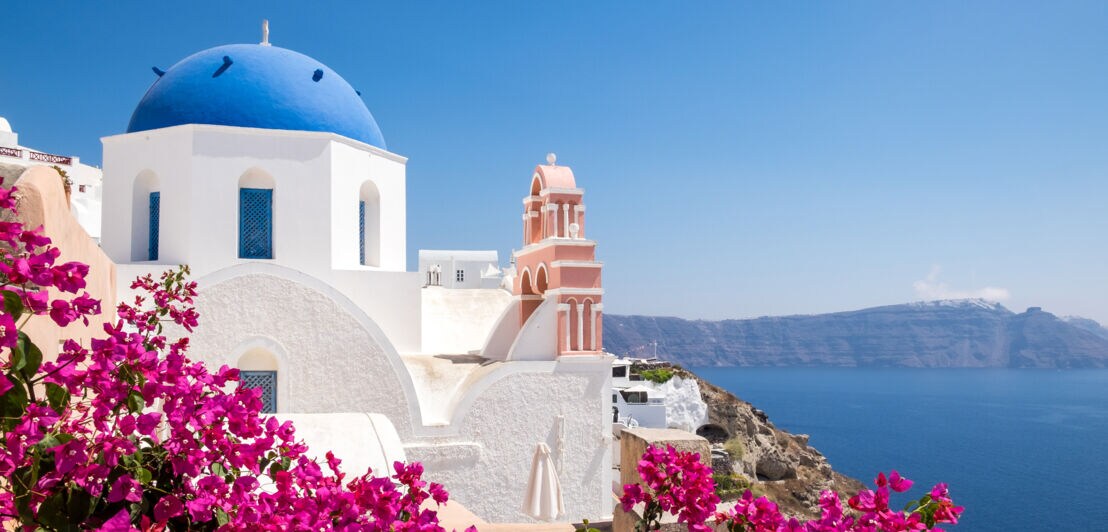 Blick auf eine weiße Kirche mit blauem Kuppeldach und pinken Bougainvillea-Blüten in Santorin, Griechenland, mit Meer und Klippen im Hintergrund