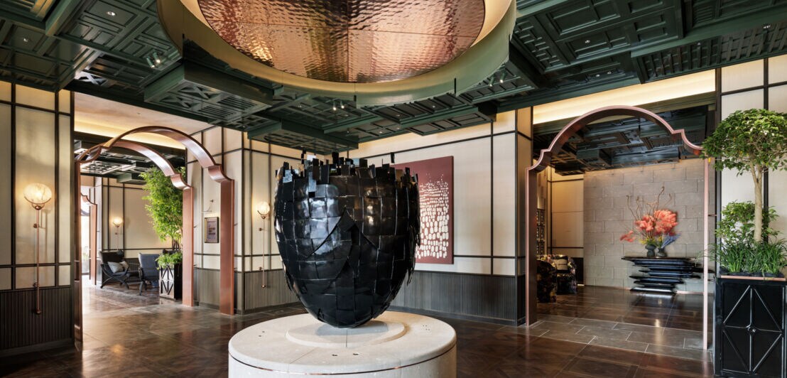 Eingangshalle mit großem, schwarzen, skulpturalen Kunstwerk auf rundem Podest, dunklem Holzparkett und dekorativer Decke