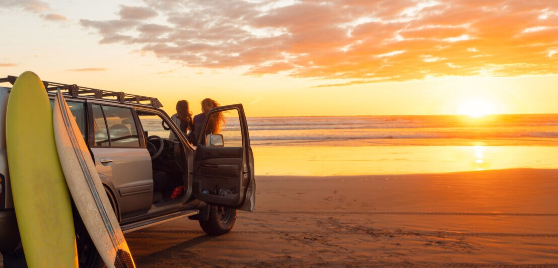 Zwei Personen stehen neben einem offenen Auto am Strand mit zwei Surfbrettern, Sonnenuntergang über dem Meer im Hintergrund