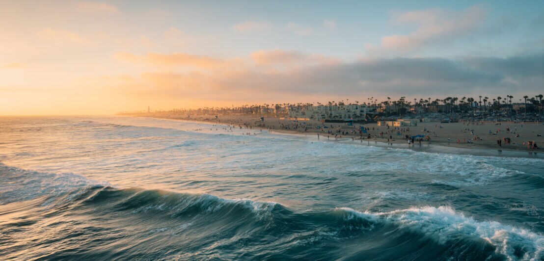 Sonnenuntergang über einem belebten Strand mit Wellen im Vordergrund und Palmen am Horizont