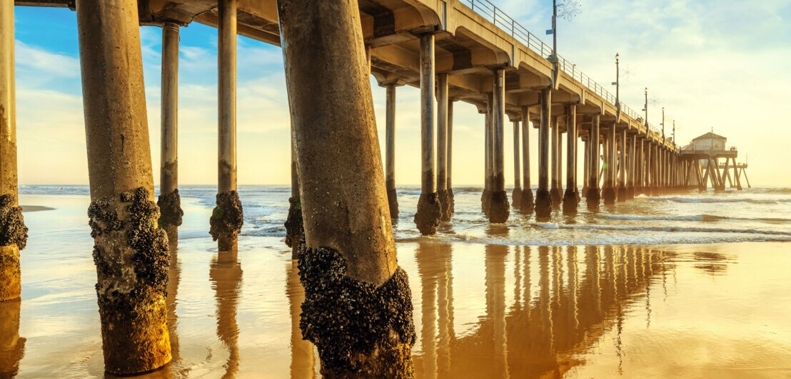 Blick unter einen langen Holzsteg mit Pfeilern im Wasser bei Sonnenuntergang am Strand