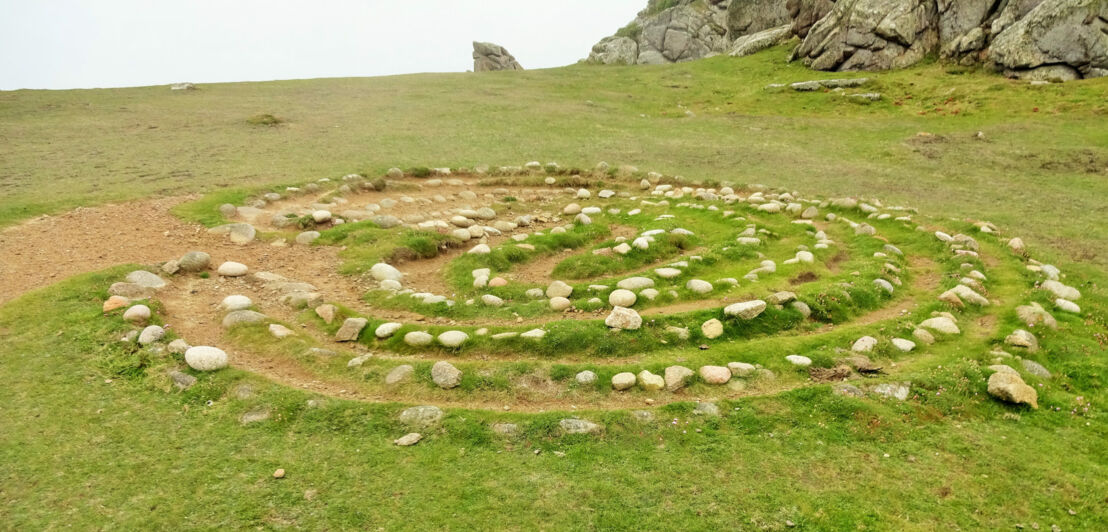 Steinspirale auf einer grasbewachsenen Fläche vor Felsen unter bewölktem Himmel