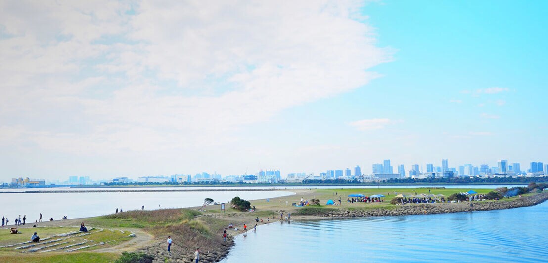 Personen auf einer künstlich angelegten Landzunge im Wasser vor der Skyline von Tokio.